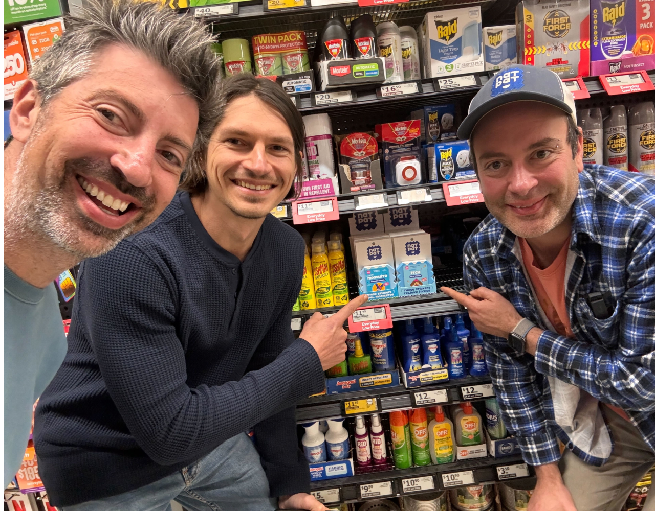 Three people pose in a supermarket aisle, pointing to NatPat products displayed on a shelf among insect repellents and household items. Product boxes are visible between them as they crouch in front of the display.