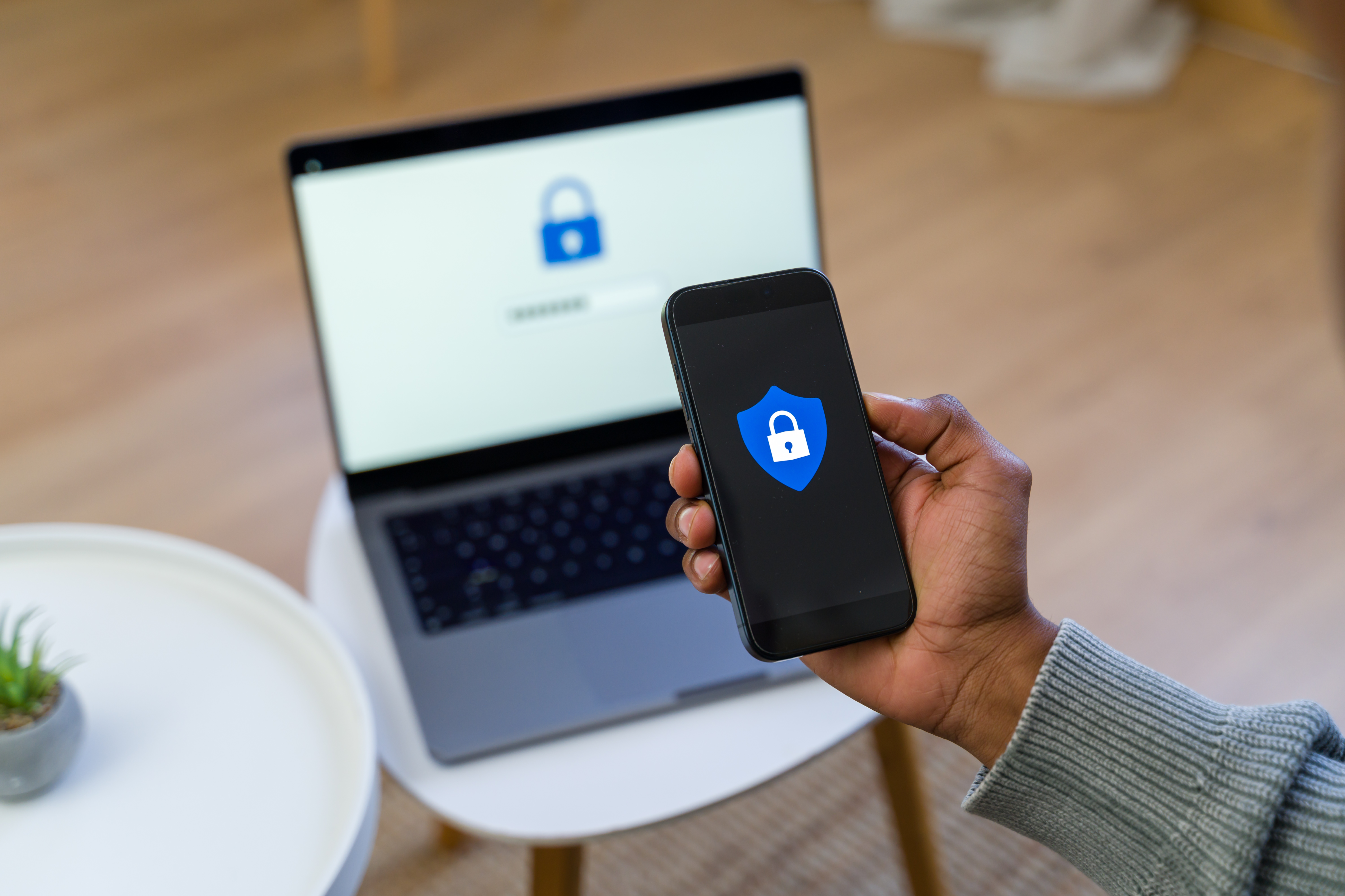 A person holds a smartphone displaying a security shield icon, with a laptop in the background, representing online safety and scam awareness.