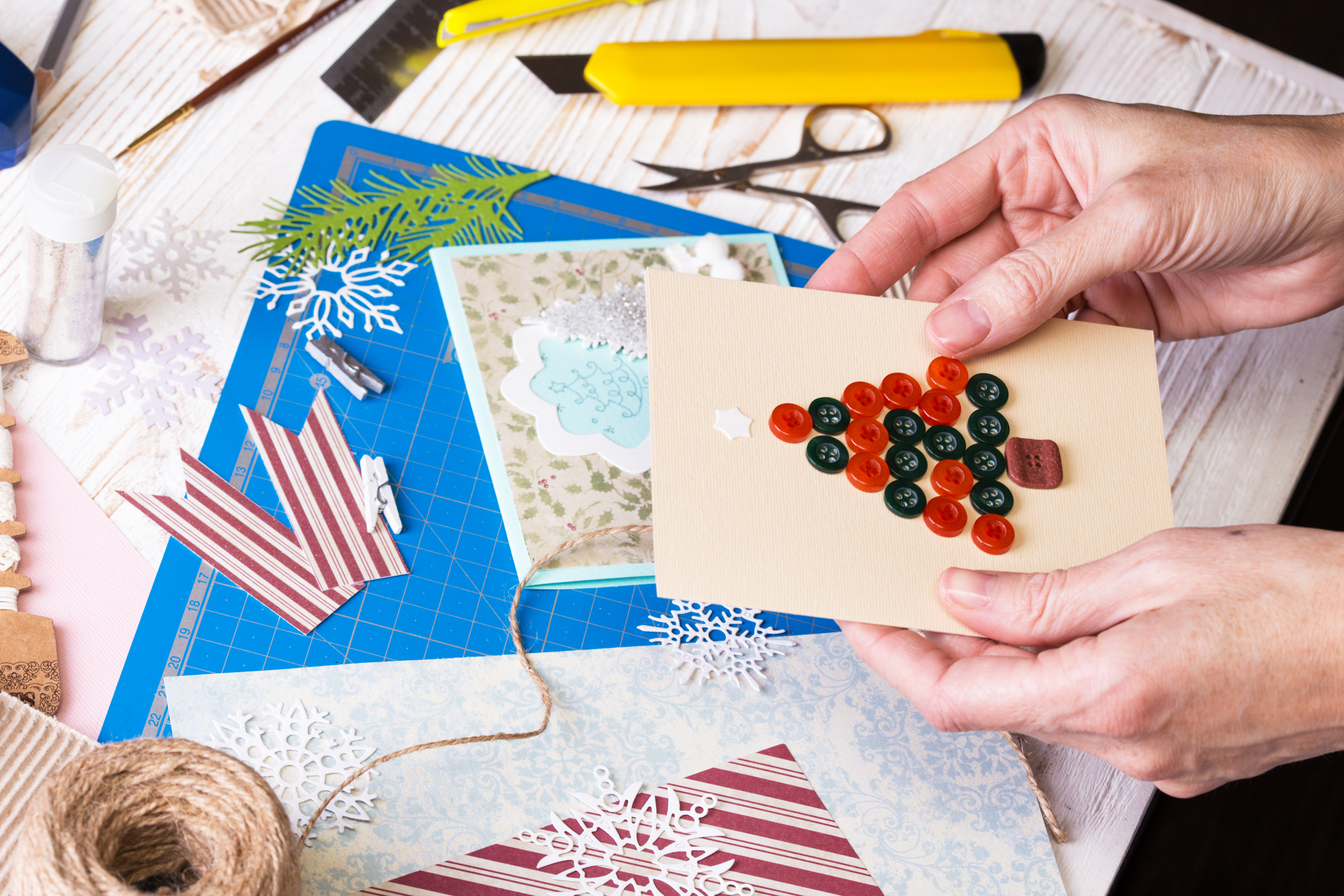 Hands hold a handmade greeting card decorated with buttons while crafting materials are arranged on a table during a card-making activity.