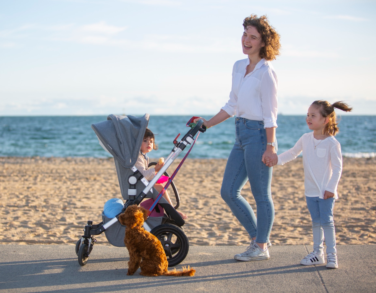 An adult walks along a beachfront promenade while pushing a stroller with a child seated inside and holding hands with another child walking beside them. A small dog sits near the stroller. The sandy beach and ocean stretch across the background under a clear sky.  Provide your feedback on BizChat