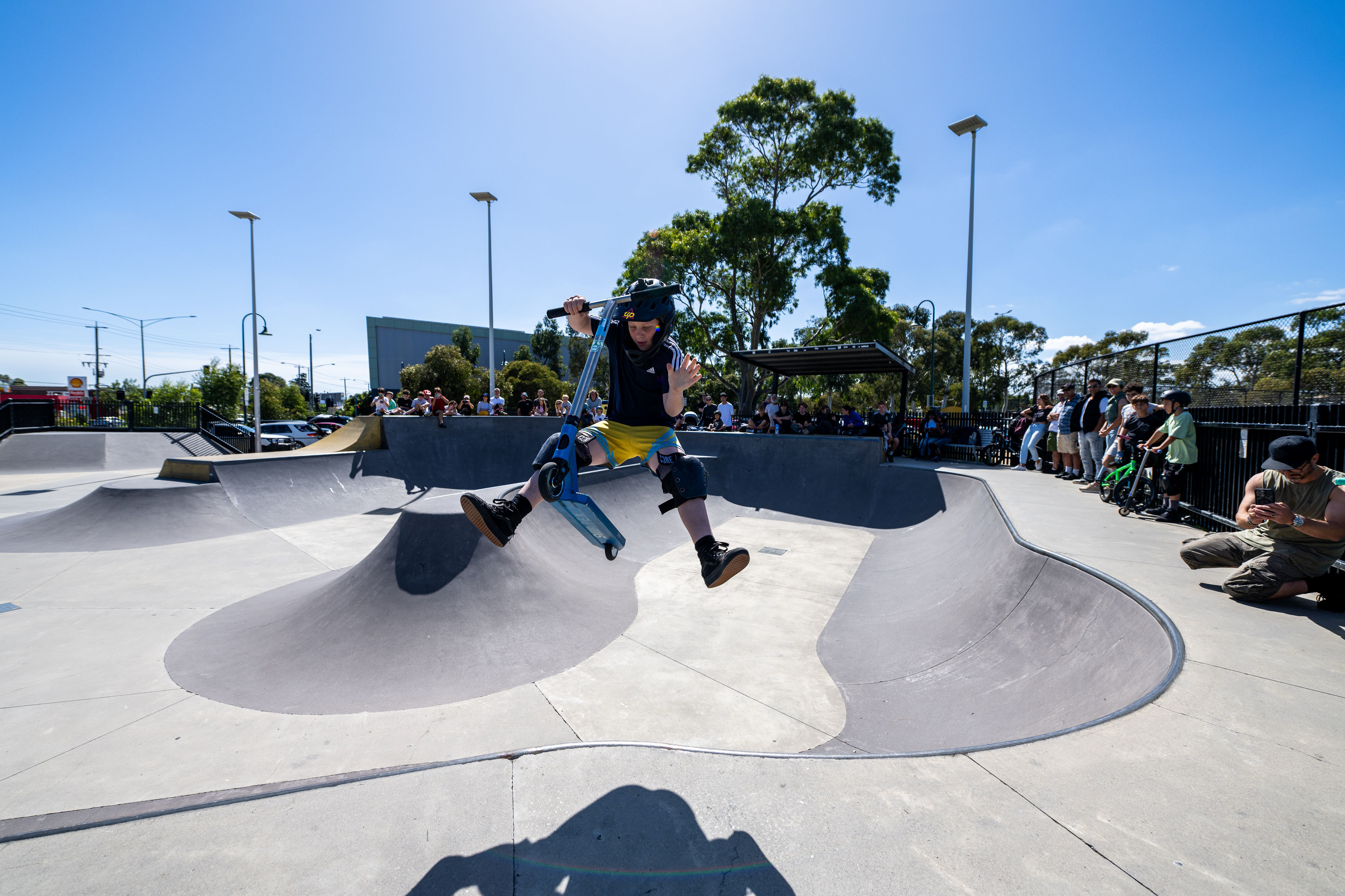 Person performing a scooter trick above a concrete bowl at a skate park while spectators watch from the sidelines.