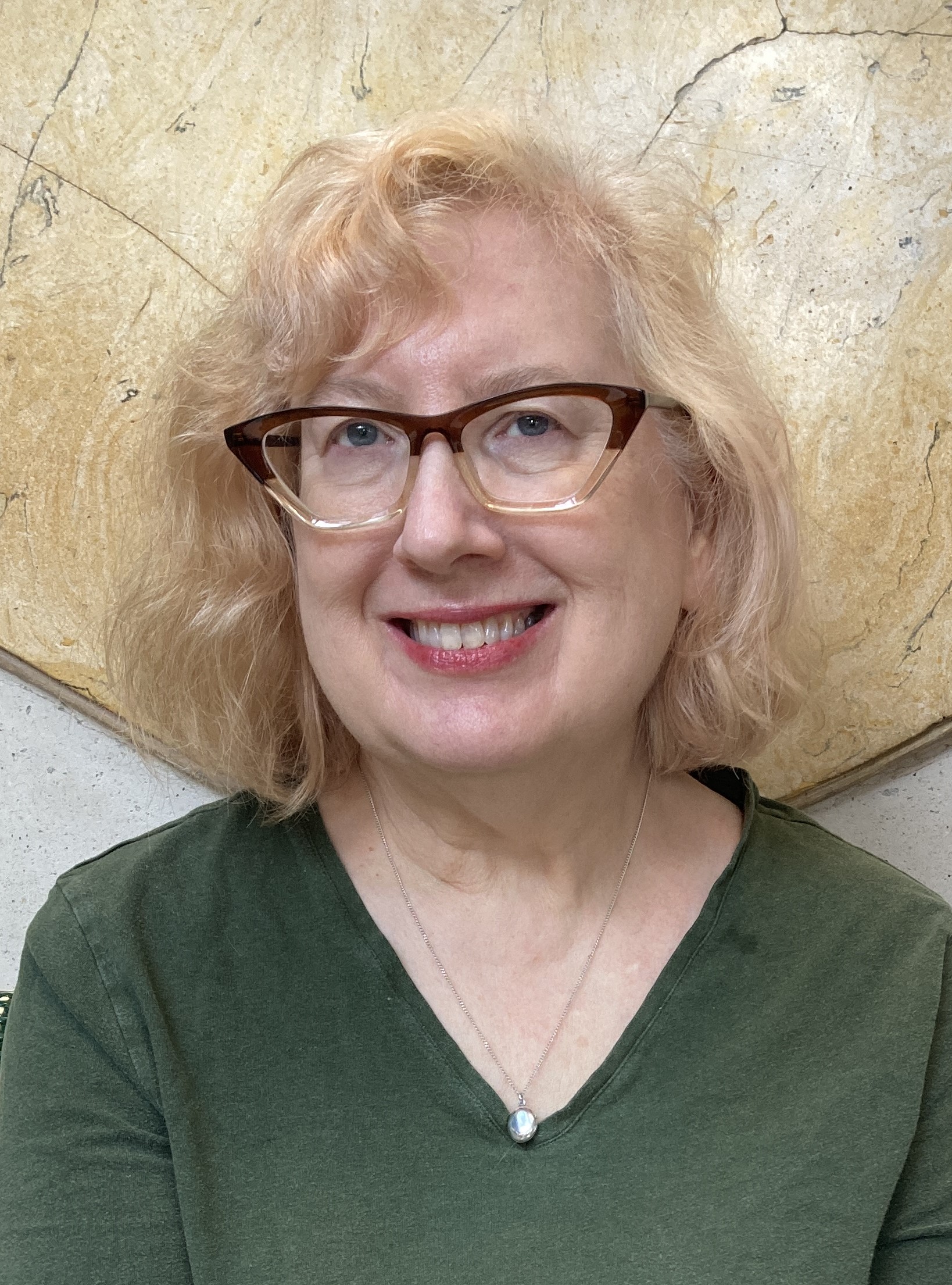 Portrait of a person wearing glasses and a green top, seated in front of a textured stone wall.