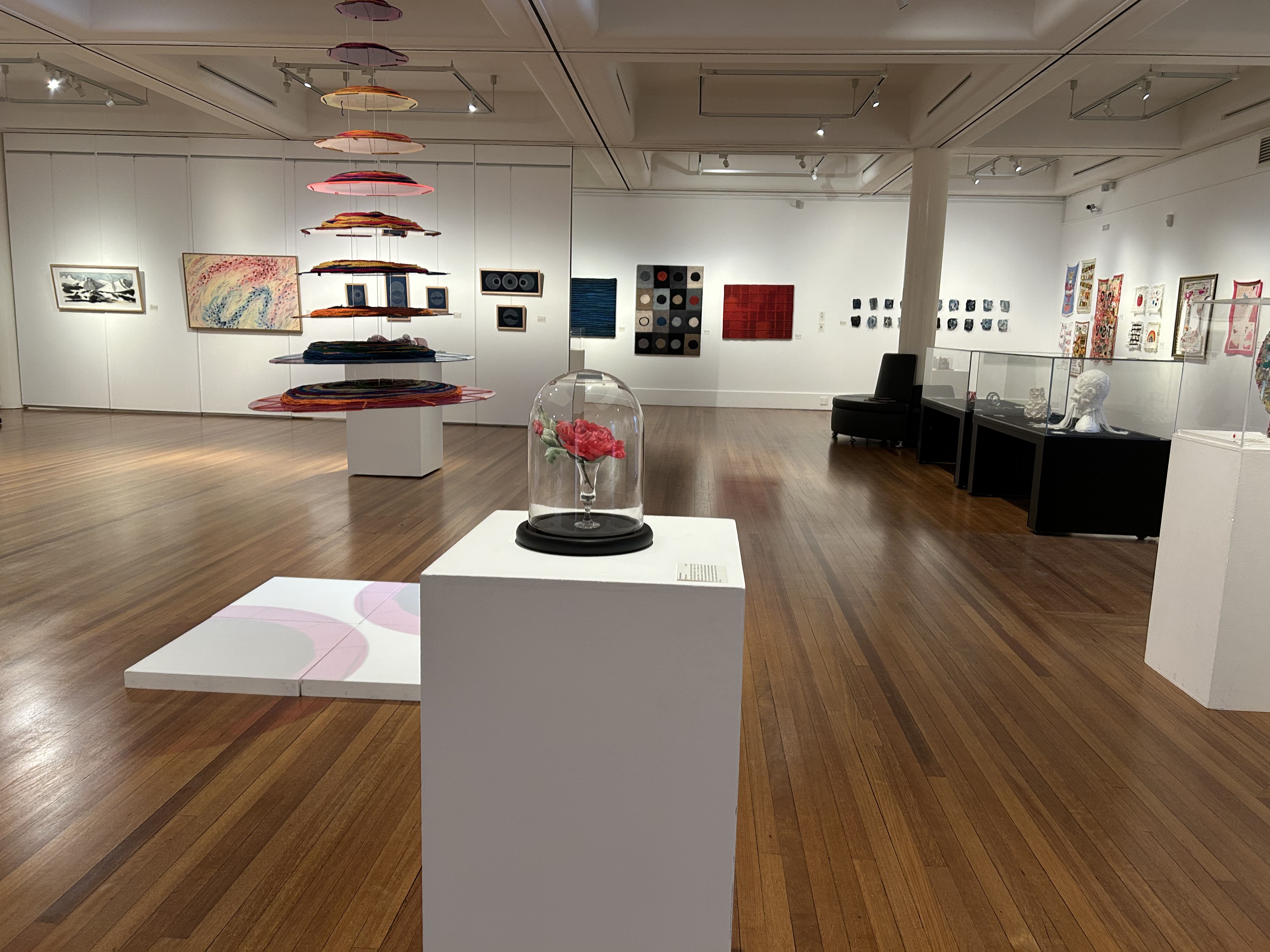 Art gallery interior with colourful textile installation suspended from the ceiling, a glass-domed vase with red flowers on a plinth in the foreground, and paintings and sculptural works displayed along white walls.