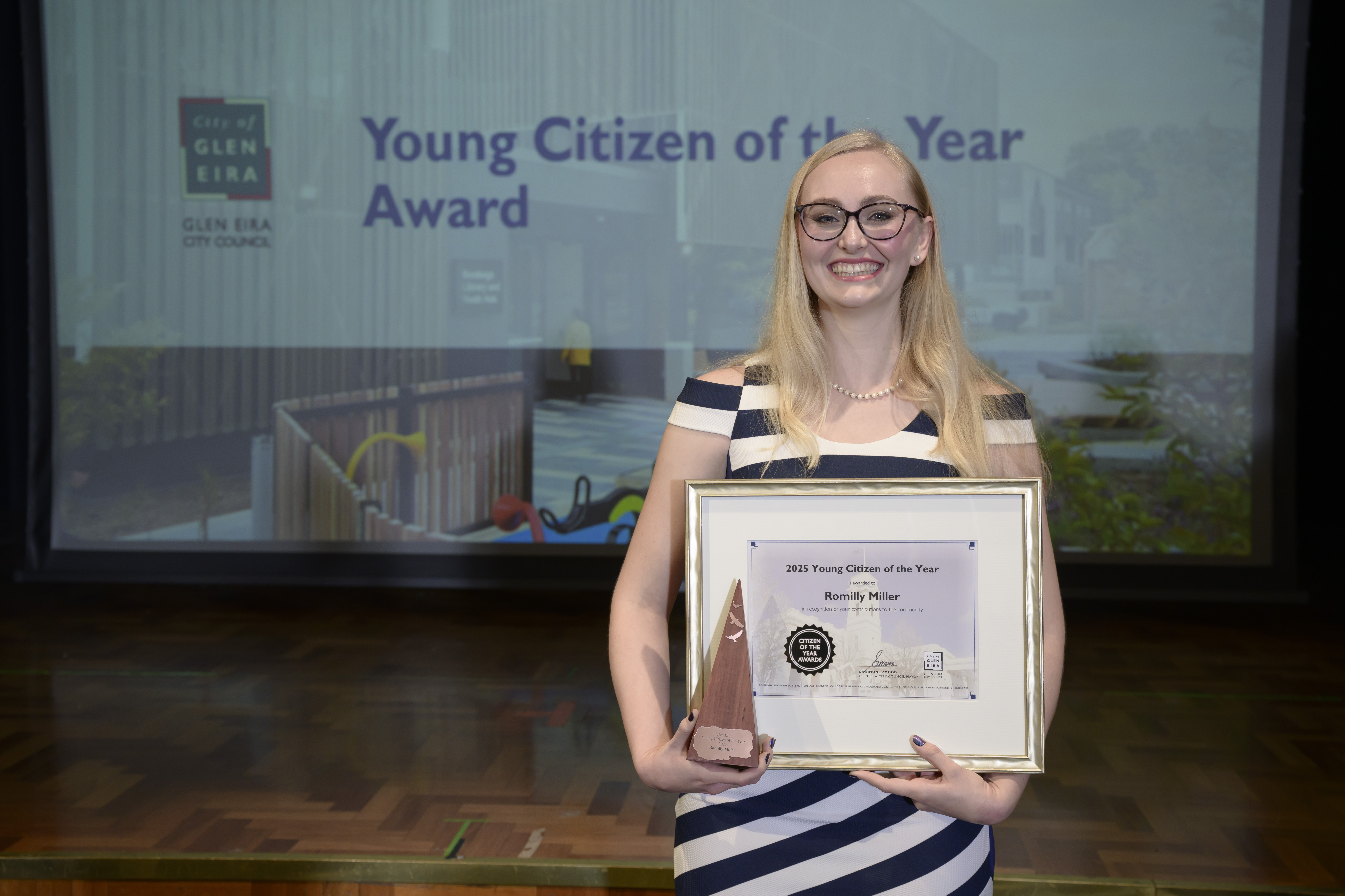 A person in a striped dress standing on a stage holding a framed certificate and a triangular award trophy. Behind them, a presentation screen shows the text “Young Citizen of the Year Award” with the City of Glen Eira branding.