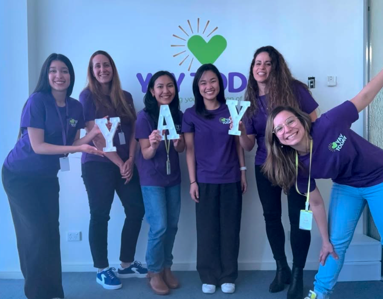 Six staff members wearing matching purple Yay Today T-shirts pose in front of a Yay Today wall logo. Three people hold large letters spelling “YAY” while another person stands with arms outstretched beside the group.